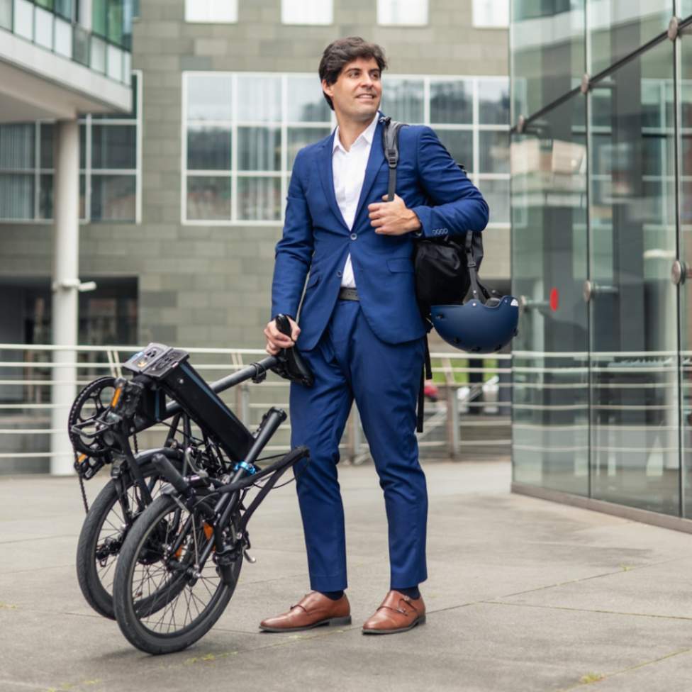Commuter in a blue suit with a blue helmet wheeling a black folded electric bike