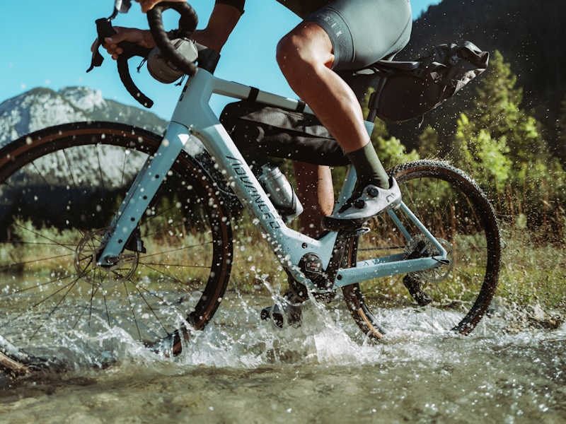 Pale blue gravel e-bike being ridden through a puddle in the mountains in sunshine